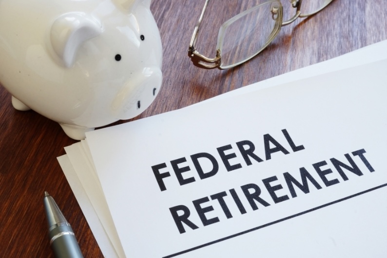 Documents reading "Federal Retirement" on a wooden desk next to a white piggy bank and glasses.