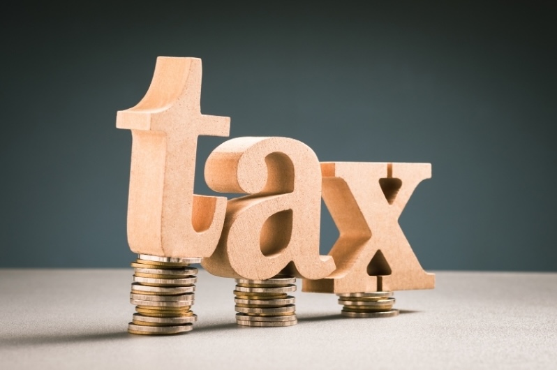 Wooden letters spelling out the word 'tax' balanced unevenly on three small stacks of coins, illustrating the complexities of the Net Investment Income Tax (NIIT)